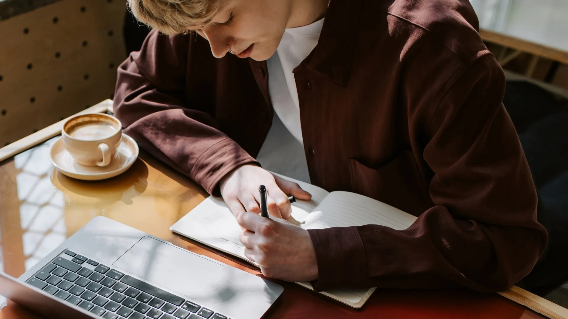 Close up of person writing in a notebook at a table with a coffee and their computer